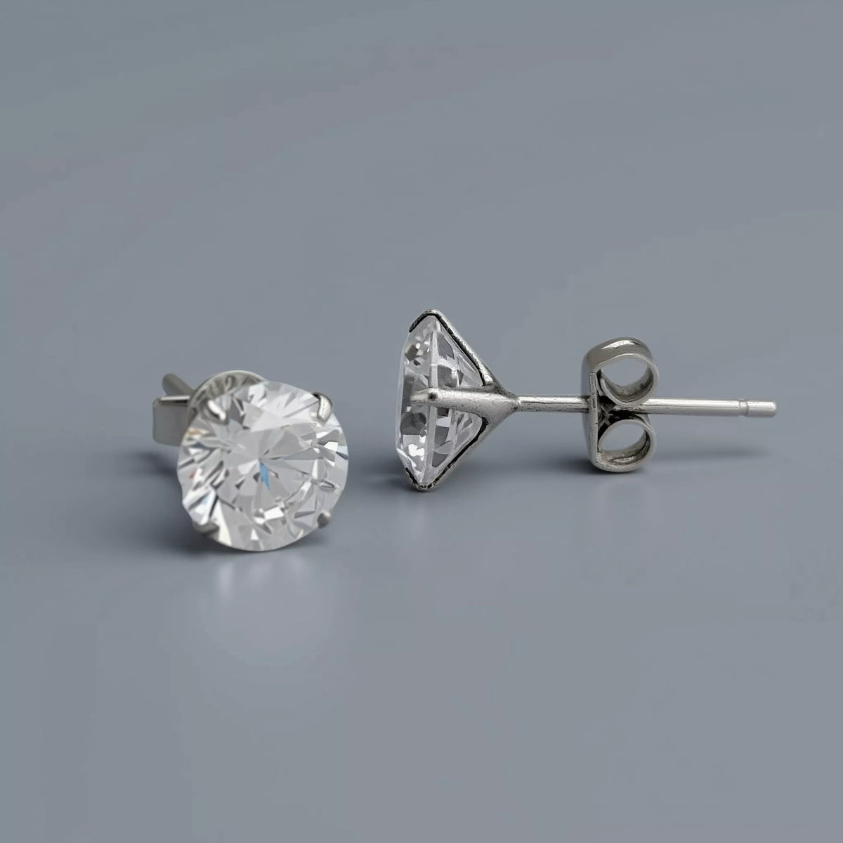 A pair of silver stud earrings crafted in 925 sterling silver featuring clear-white round cubic zirconia gemstones, and showing a triangular prong setting with a butterfly back, displayed on a smooth cool mid grey background with soft diffused lighting.
