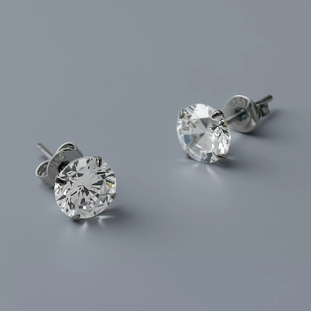 A pair of silver stud earrings crafted in 925 sterling silver featuring clear white round cubic zirconia stones displayed on a smooth cool mid grey background with soft diffused lighting. One earring faces forward and the other angled to the side.
